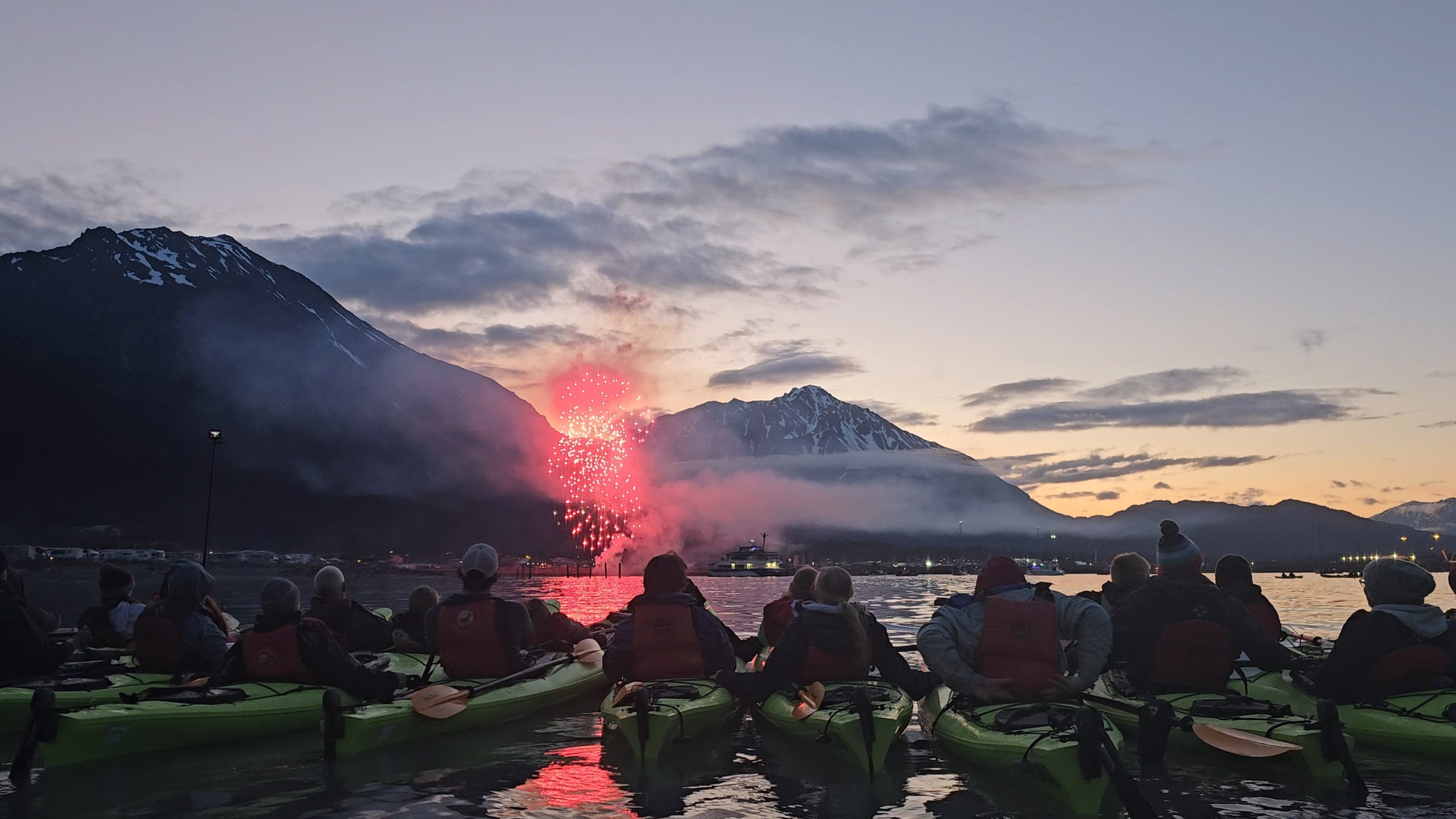 People in kayaks watching fireworks over a mountain at sunset.