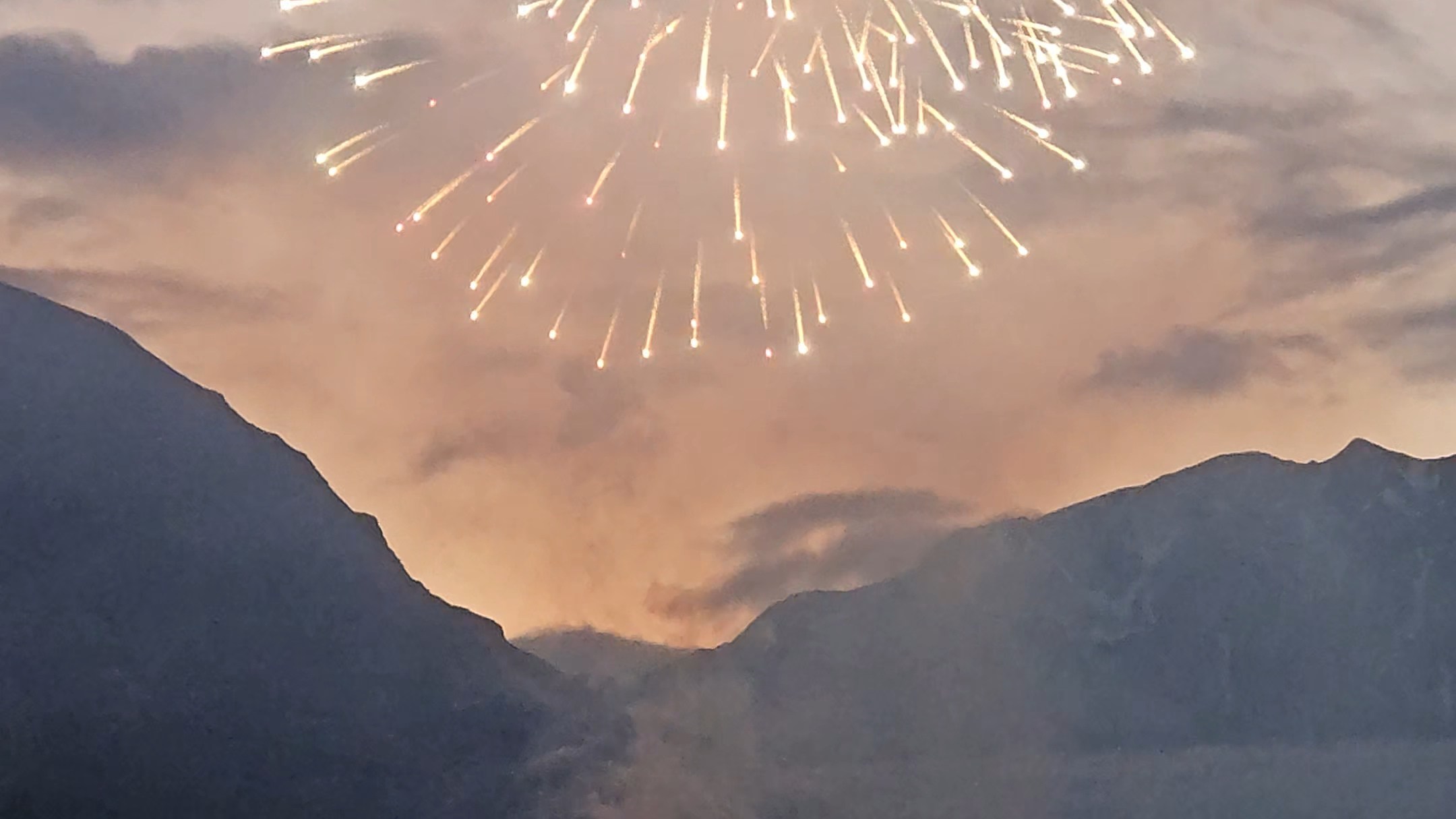 Fireworks burst over mountains at dusk with people watching and taking photos.