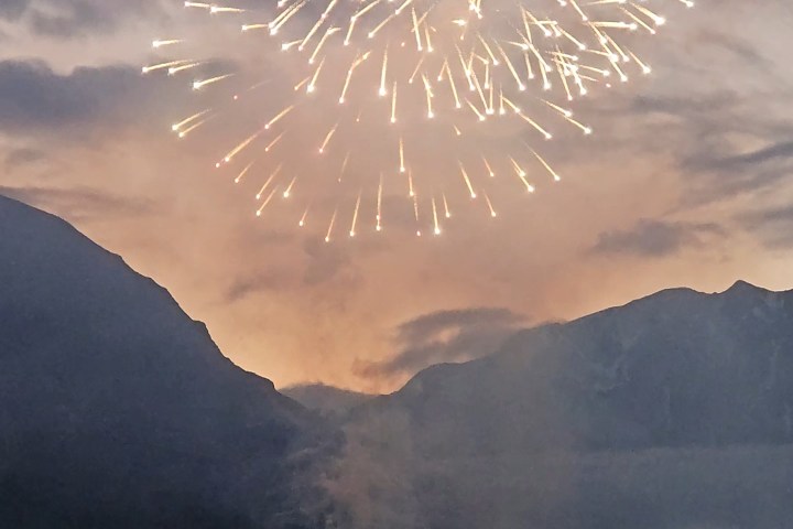 Fireworks burst over mountains at dusk with people watching and taking photos.