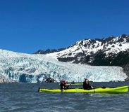 Two people kayaking on a lake with a snow-capped mountain and glacier in the background.