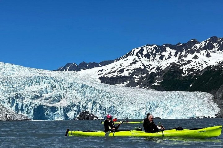 Two people kayaking on a lake with a snow-capped mountain and glacier in the background.