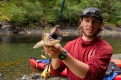Person holding a fish on a fishing rod by a riverbank, wearing a red shirt and cap.