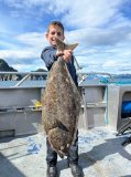 Child holding a large fish on a boat with mountains in the background under a cloudy sky.