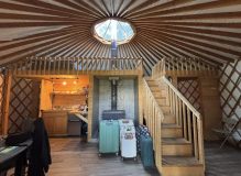 Interior of a yurt with a round skylight, wooden stairs, stove, and kitchen area.