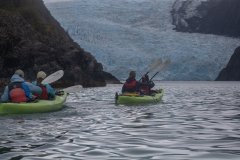 Kayakers on calm water near a glacier under cloudy skies.