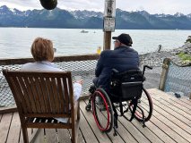 Two people sit on a deck, one in a wheelchair, overlooking a lake with mountains in the background.