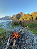 Campfire by a lake with a person taking photos of mountains in the distance.