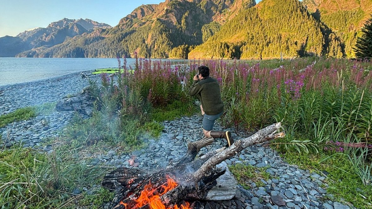 Campfire by a lake with a person taking photos of mountains in the distance.