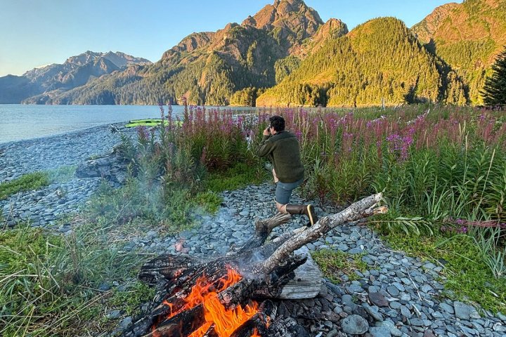 Campfire by a lake with a person taking photos of mountains in the distance.