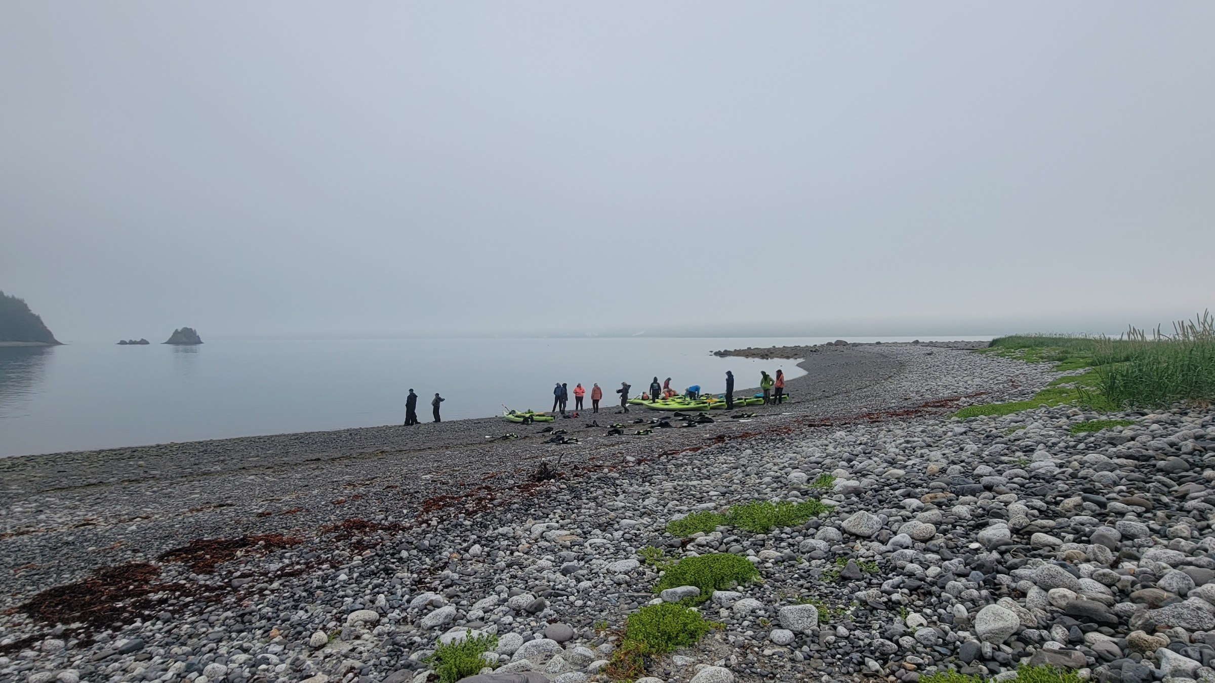 People with kayaks on a rocky beach beside calm foggy water.