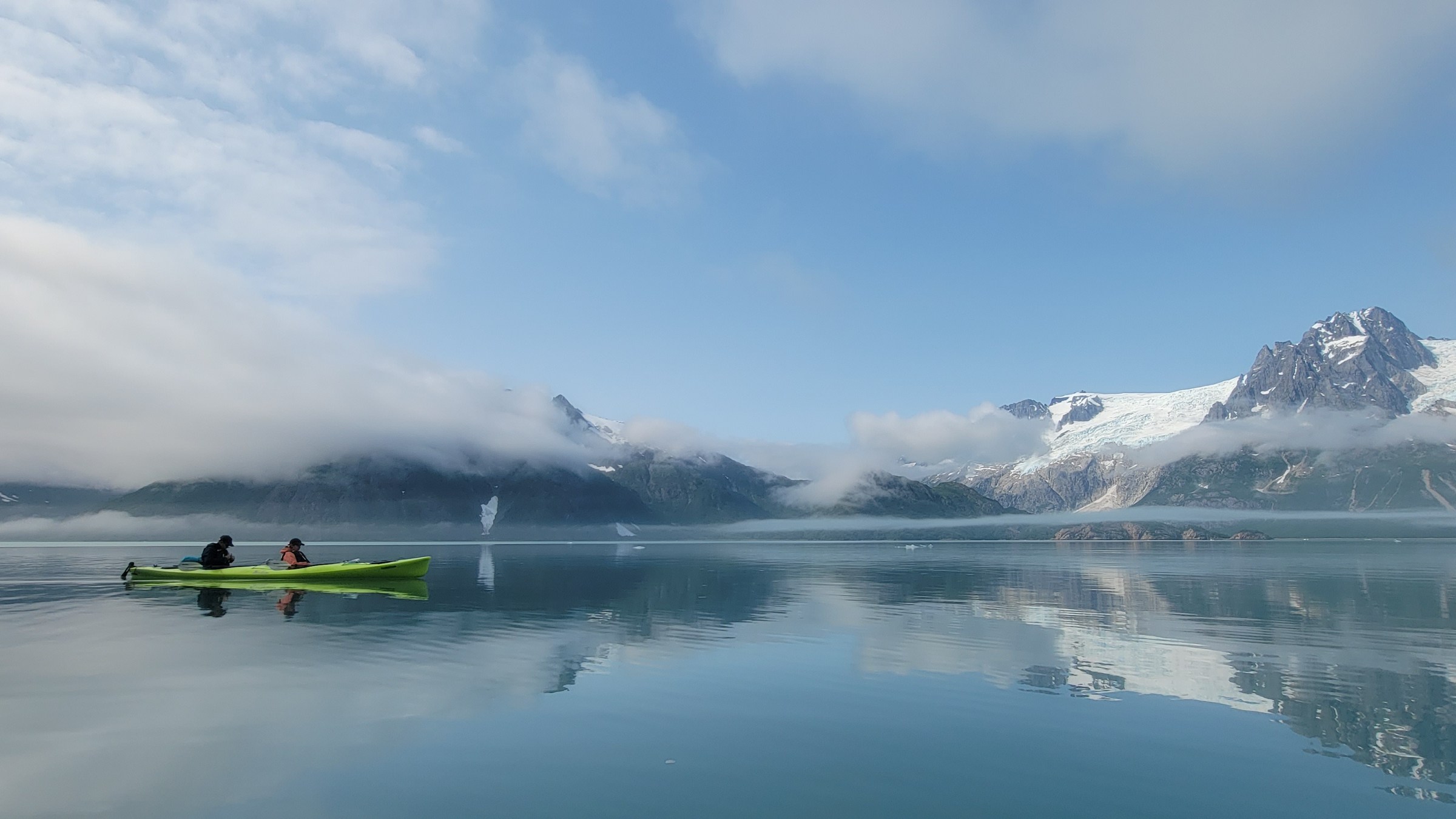 Two people in a green kayak on a calm lake beside snowy mountains under a blue sky.