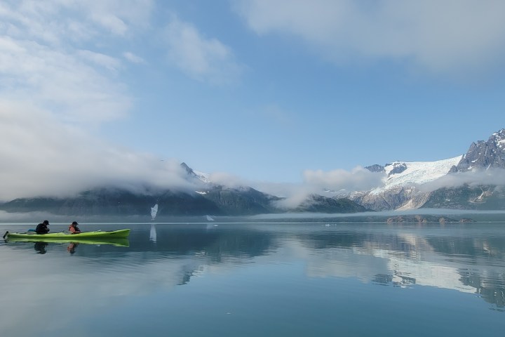 Two people in a green kayak on a calm lake beside snowy mountains under a blue sky.