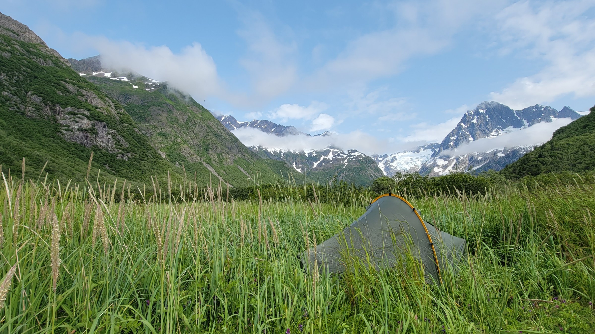 Tent in grassy field with mountains and clouds in background.