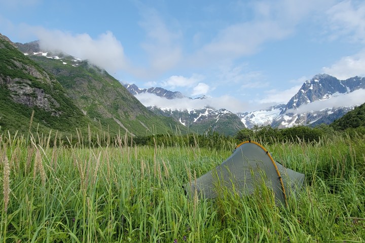 Tent in grassy field with mountains and clouds in background.