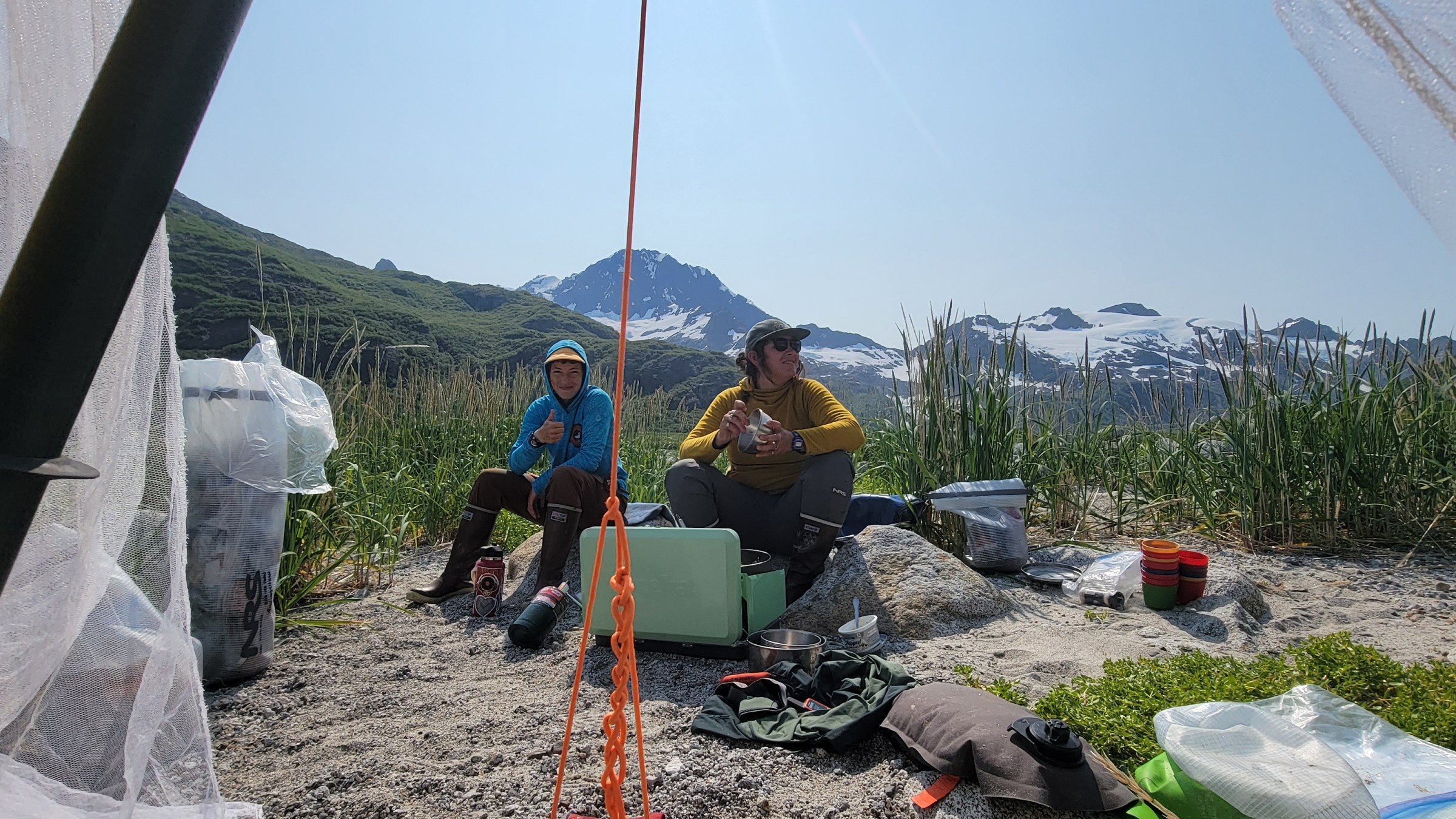 Two people camping by a lake, surrounded by mountains and gear; clear blue sky above.