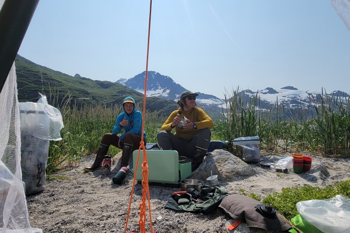 Two people camping by a lake, surrounded by mountains and gear; clear blue sky above.
