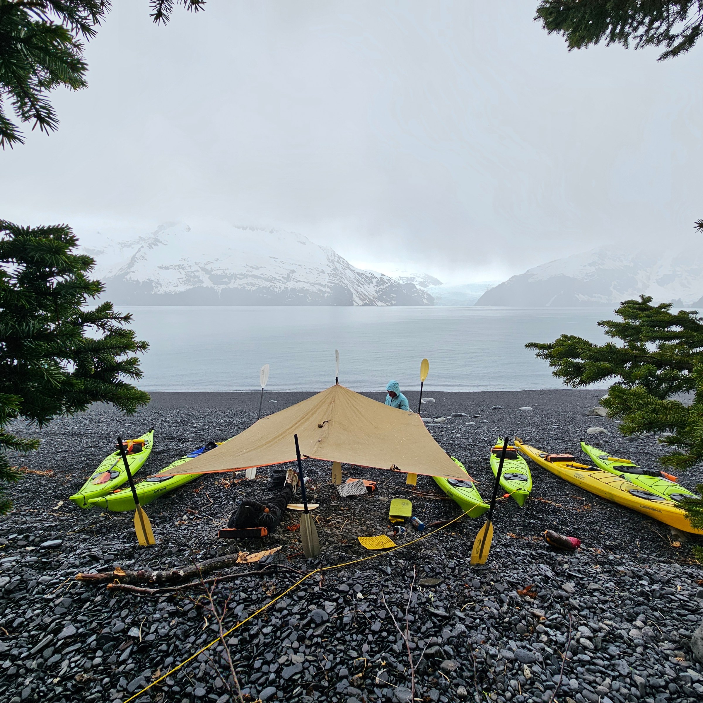Tarp shelter on rocky beach with kayaks, snowy mountains in the background.