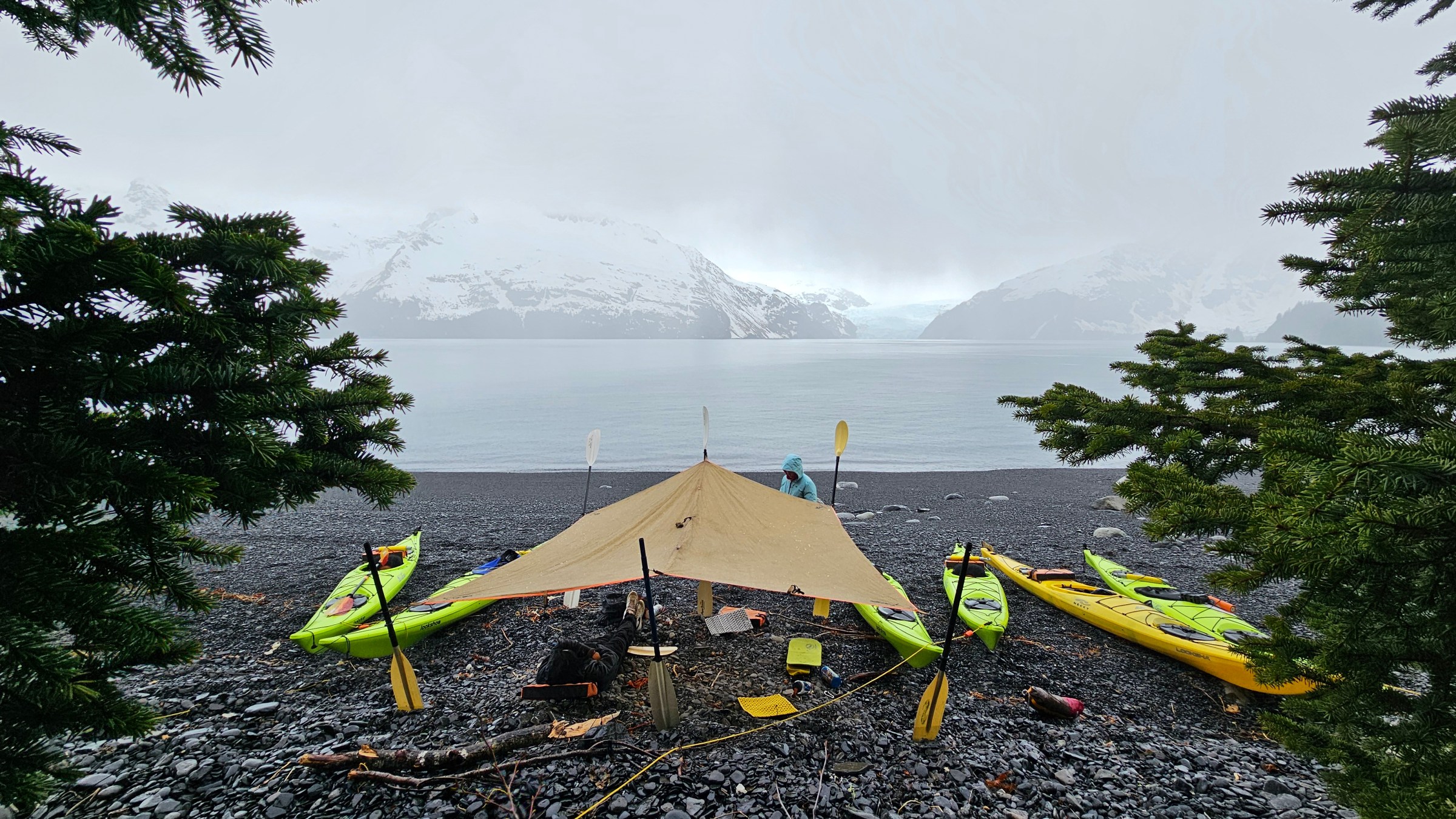 Tarp shelter on rocky beach with kayaks, snowy mountains in the background.