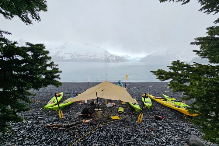 Tarp shelter on rocky beach with kayaks, snowy mountains in the background.