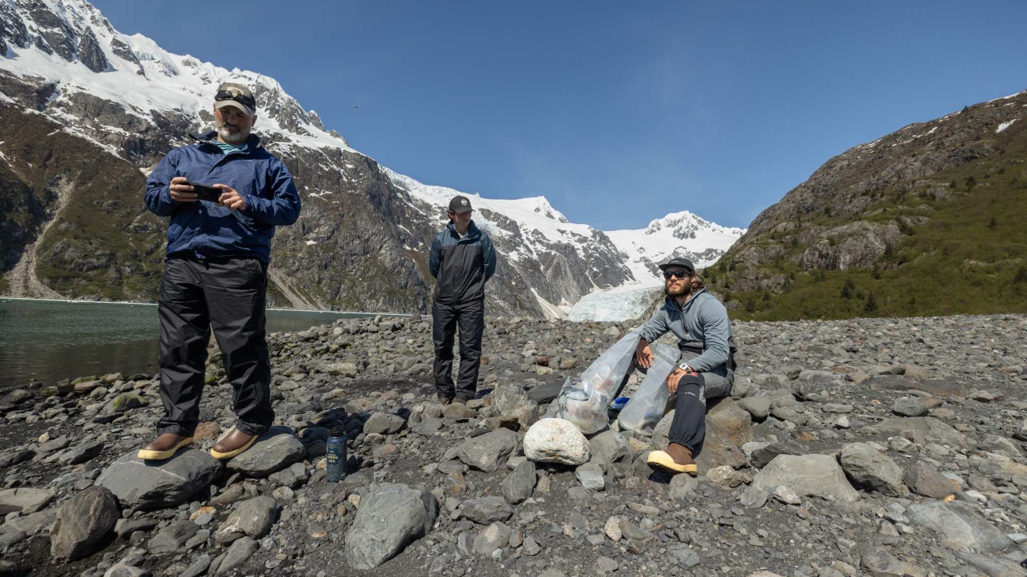 Three people on rocky terrain by snowy mountains, two standing, one sitting with bags, under clear blue sky.