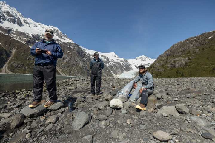 Three people on rocky terrain by snowy mountains, two standing, one sitting with bags, under clear blue sky.