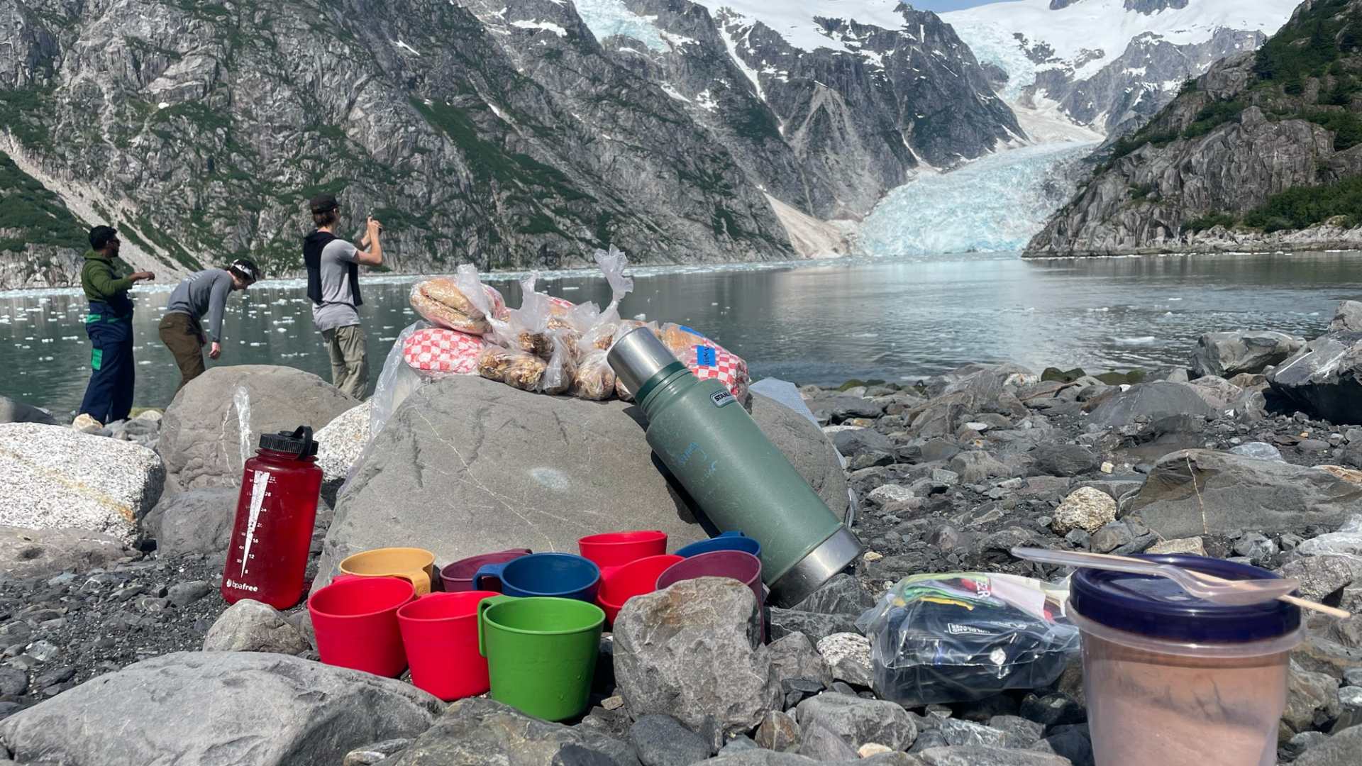 Picnic setup with cups and snacks on rocky shore, glacier and mountains in background.