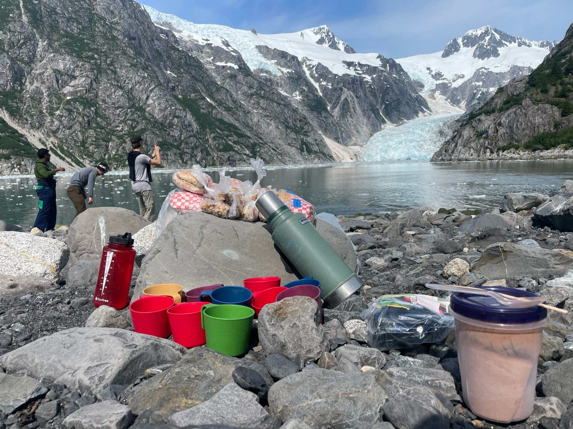 Picnic setup with cups and snacks on rocky shore, glacier and mountains in background.
