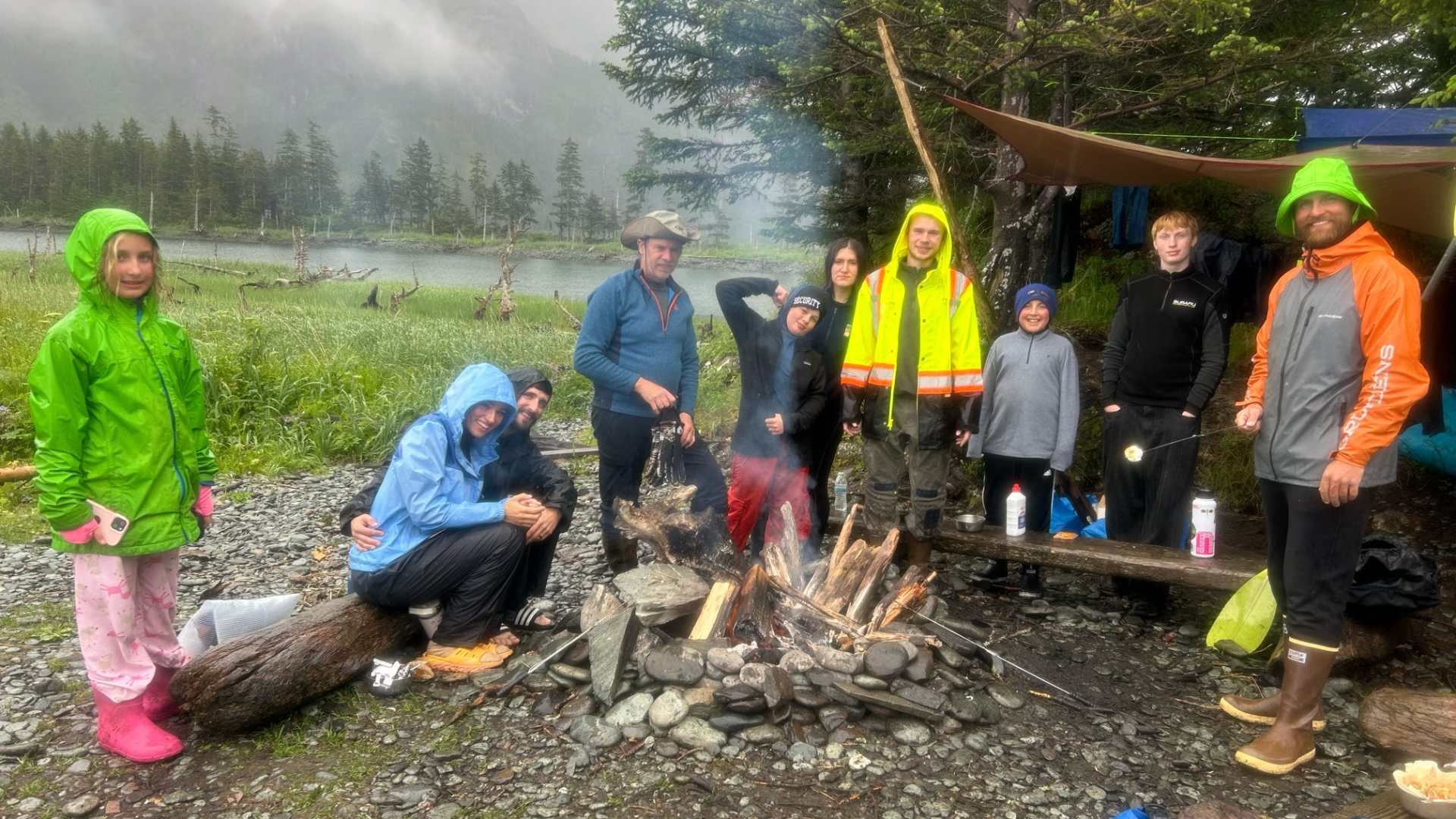 Group of people in raincoats around a campfire in a forested area on a cloudy day.