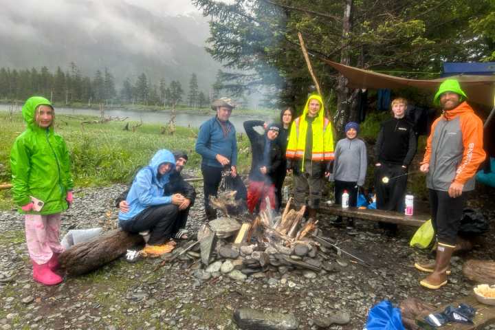 Group of people in raincoats around a campfire in a forested area on a cloudy day.
