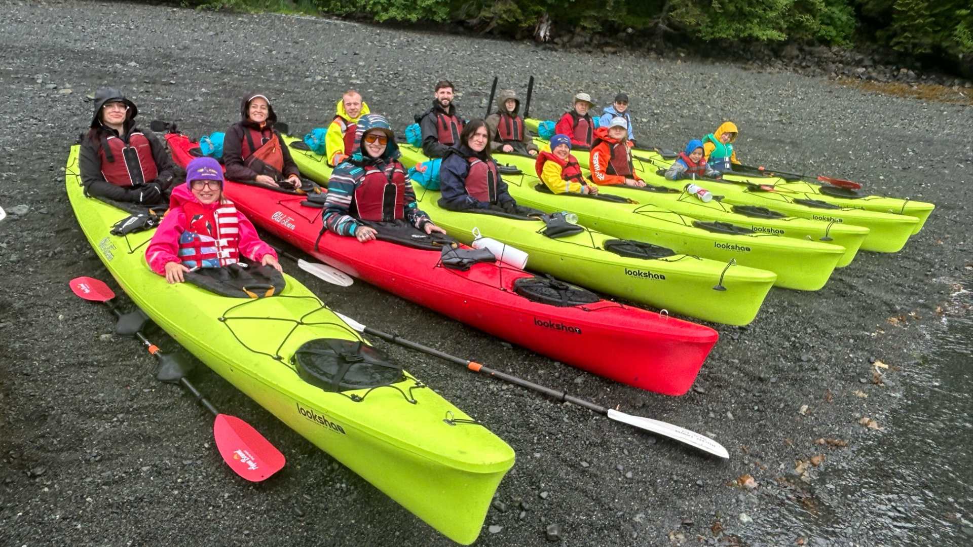 Group of people in life vests sitting in kayaks on a pebbly shore with green trees behind them.