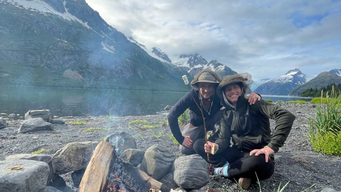 Campfire with people sitting nearby, holding a s'more, mountains in the background.