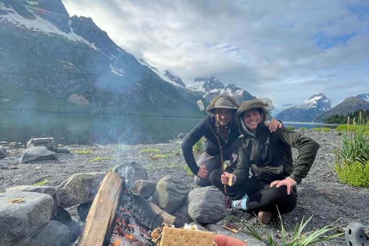 Campfire with people sitting nearby, holding a s'more, mountains in the background.