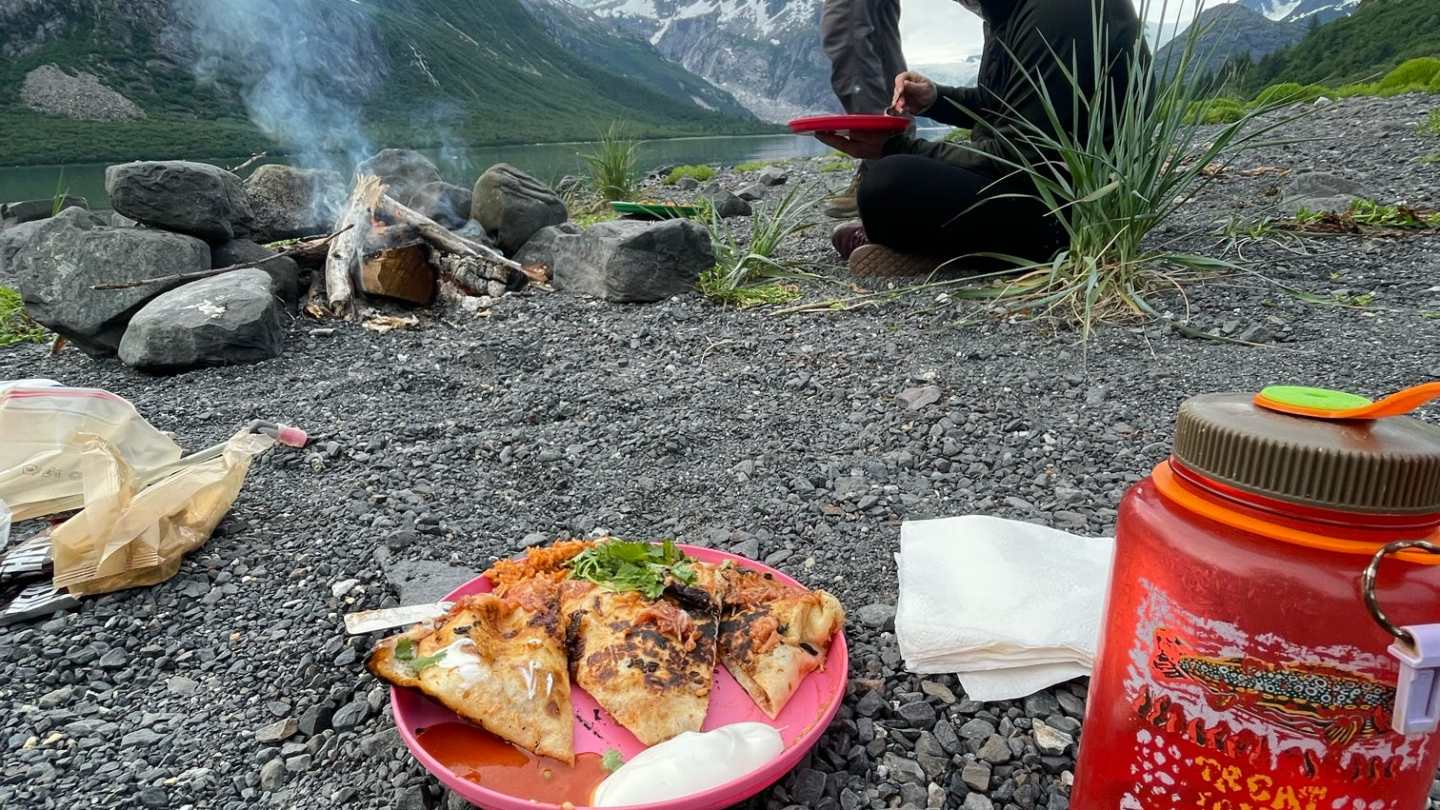 Two people by a campfire on rocky ground, with mountains, pizza on a plate, and a red water bottle nearby.