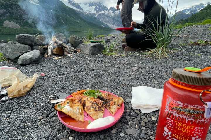 Two people by a campfire on rocky ground, with mountains, pizza on a plate, and a red water bottle nearby.