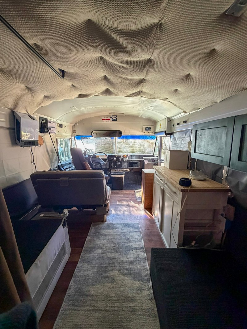 Interior of a converted bus with seating, carpet, and cabinetry; view towards the driver's seat.